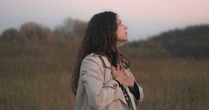 A photo of a woman standing in a field with her hands over her heart.